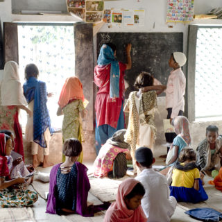 A teacher with a large group of students in an under-resourced classroom with crumbling walls. Students are sitting on the ground while the teacher writes on a chalkboard.