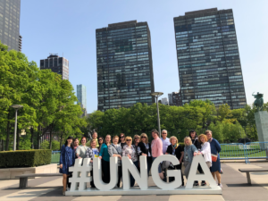 Group of people smiling while standing behind a large sign that reads "#UNGA". Large city buildings in background.