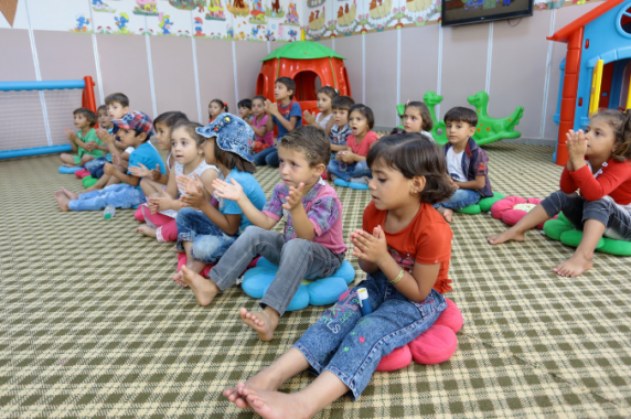 Young children who are refugees sitting on the floor of a school, repeating a song while clapping hands