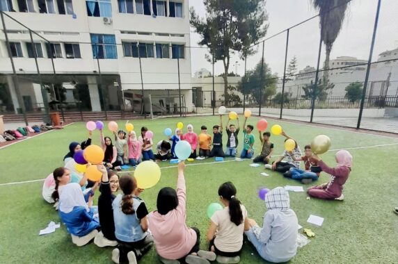 Photo of kids sitting in a schoolyard in a circle, holding balloons