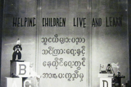 A black-and-white historical photo of a sign that says "Helping children live and learn" in English at the top and has words underneath in Burmese. Children's play blocks and other toys are in front of it.