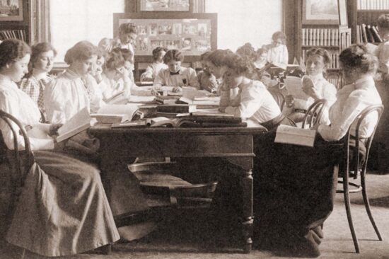 Black-and-white historical photo of teachers in dresses, sitting around a table, collaborating.