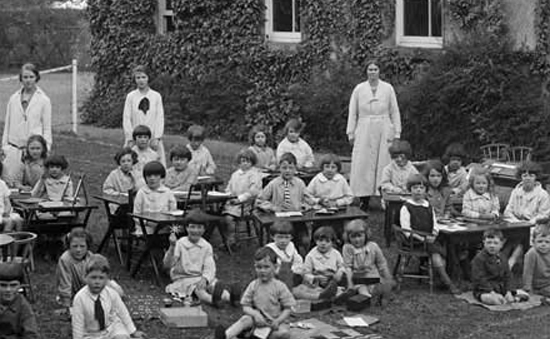 A black-and-white historical photo of teachers outdoors with students sitting in desks on a lawn.