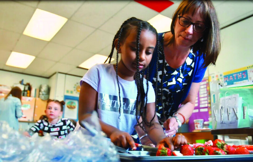 Photo of a teacher helping a student cut strawberries in a classroom
