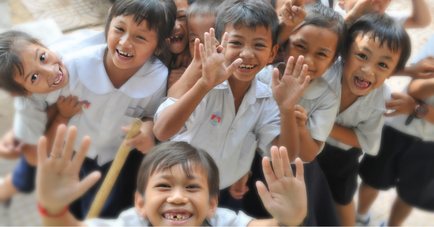 A group of smiling young children in school unforms, holding up their hands to wave