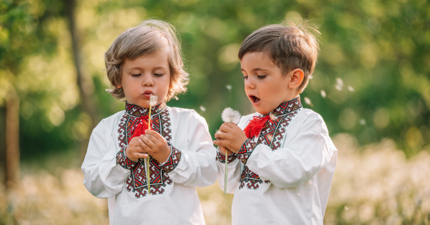 Two young Ukrainian children wearing traditional shirts in a field, blowing on dried dandelions.