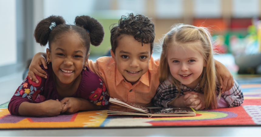 Photo of three smiling children laying on the floor with a book they are reading