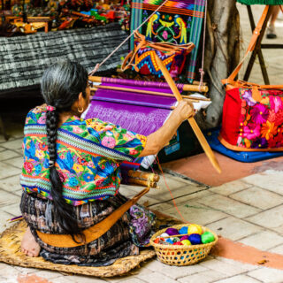 A woman in Guatalmala, sitting on a mat on the floor, weaving a traditional tapestry with bright colors.