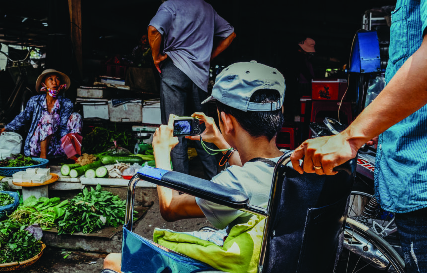 A boy sitting in a wheelchair at a market, holding a digital camera and taking a photo of a woman selling fresh green vegetables