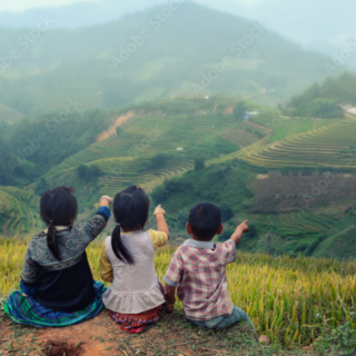 Three children sitting on a hill with green fields around, pointing in the distance.