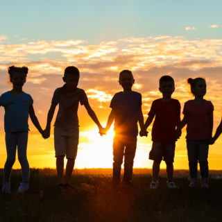 Shadows of five children holding hands as the sun sets in the distance.