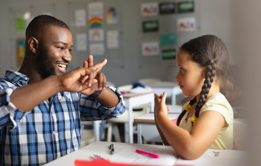 A teaching and young student sitting at a desk together, smiling and using sign language.