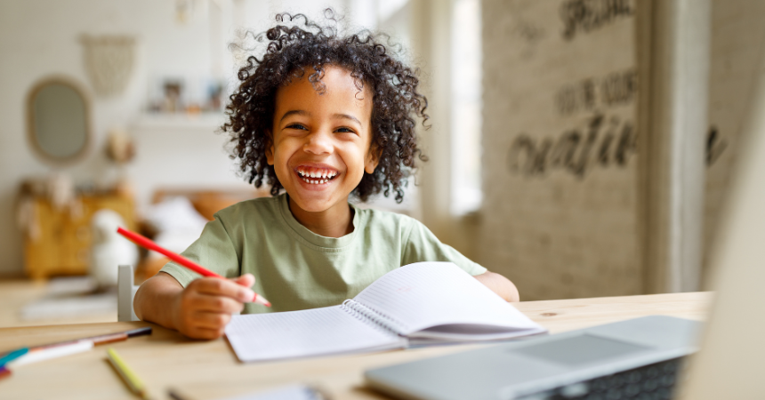 A young boy happily sitting at a desk holding a pencil and writing in a notebook.