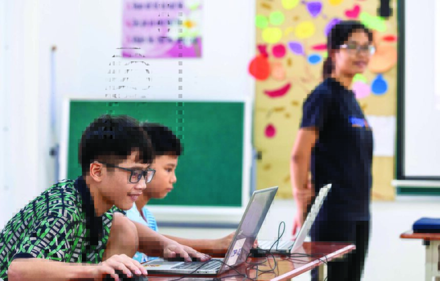 Two young students using laptops in a classroom, with a teacher students near them.