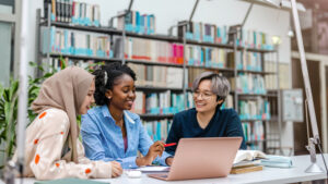 Three young people in a library, excitedly looking at and discussing a post on a laptop.