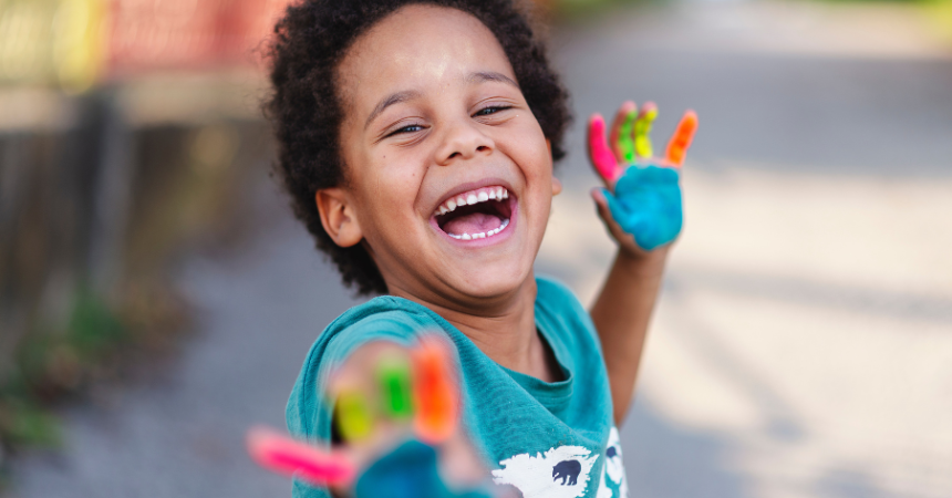 A happy young child outdoors, smiling and laughing, with multicolored painted hands, representing creativity featured in the childhood education magazine.