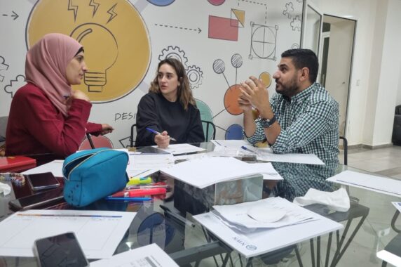 Three teachers sitting together in a classroom, talking and sharing their experiences.