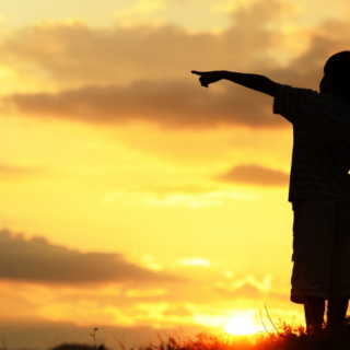 Two children standing together at sunset, with one of them pointing to something in the distance to show something to the other.