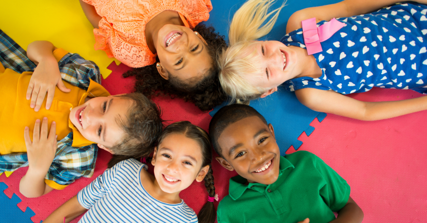 Five smiling children lying together in a circle on a colorful mat, smiling.